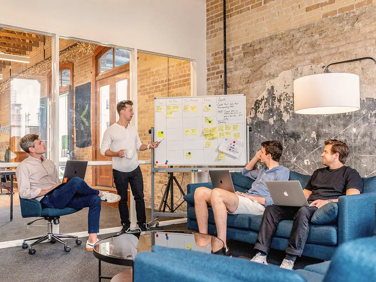 A group of young professionals sitting in an office setting, chatting casually and reviewing a whiteboard.
